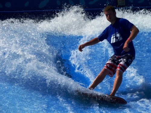 A surfer rides a wave on a blue artificial surf simulator, splashing water around him.