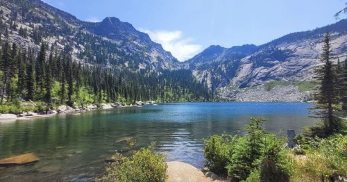 A serene mountain lake surrounded by lush greenery and towering peaks under a clear blue sky.