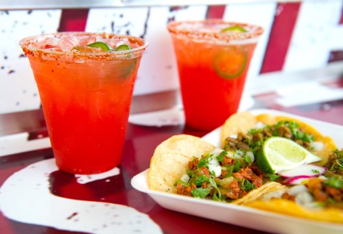 Two colorful drinks with lime slices beside a plate of tacos topped with onions and cilantro.