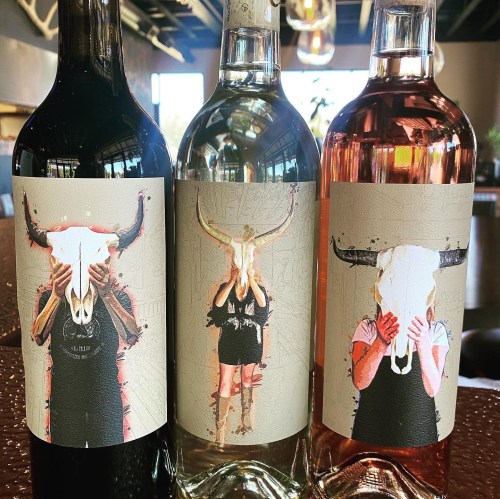 Three wine bottles with artistic labels featuring a person holding a bull skull, in red, white, and rosé varieties.