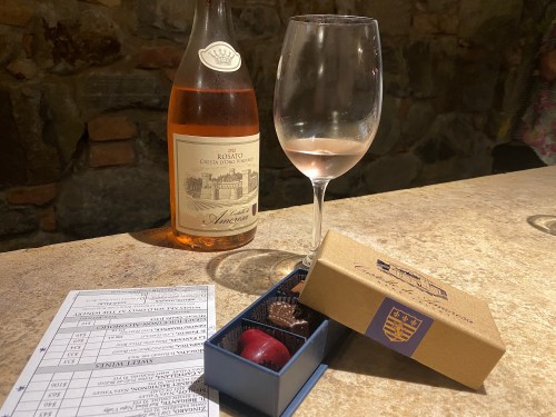 A bottle of rosé wine, a glass of wine, and a box of chocolates on a stone countertop with a tasting menu.