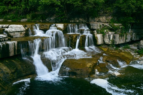 A serene waterfall cascading over rocky ledges into a calm pool, surrounded by lush greenery.