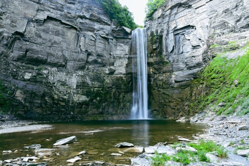 A tall waterfall cascades down rocky cliffs into a serene pool surrounded by lush greenery.