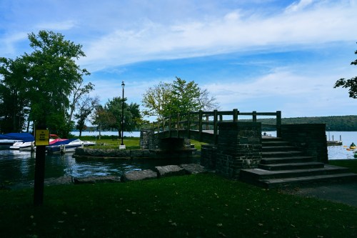 A serene lakeside scene featuring a small bridge, grassy area, and boats on the water under a blue sky.
