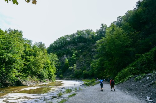 Two hikers walk along a gravel path beside a river, surrounded by lush green trees and a rocky hillside.