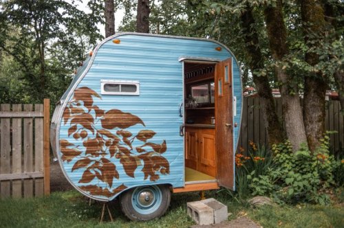 A vintage blue trailer with wooden accents, surrounded by trees and flowers, parked beside a wooden fence.