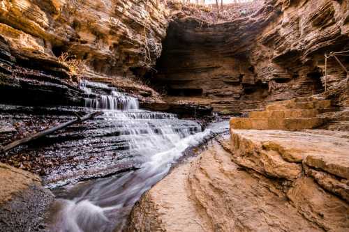 A serene waterfall cascades over rocky steps in a natural canyon, surrounded by textured stone walls and autumn foliage.