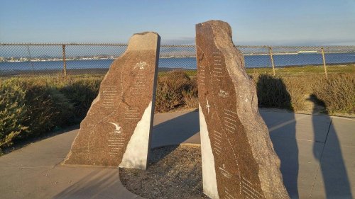 Two large stone monuments with engravings, set against a scenic waterfront view and a clear sky.