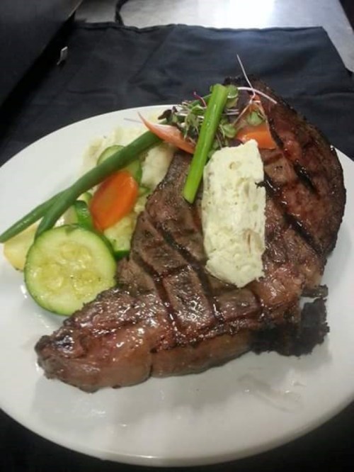 A grilled steak on a plate, topped with herb butter, accompanied by vegetables and mashed potatoes.