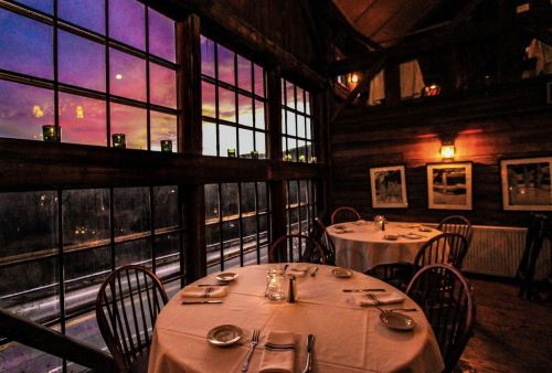 Cozy restaurant interior with tables set for dining, large windows showcasing a vibrant sunset.