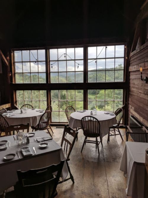 Cozy restaurant interior with round tables, wooden chairs, and large windows showcasing a scenic view of greenery.