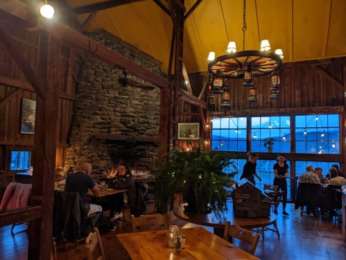 Cozy restaurant interior with wooden beams, stone fireplace, and warm lighting, featuring diners and a scenic view outside.
