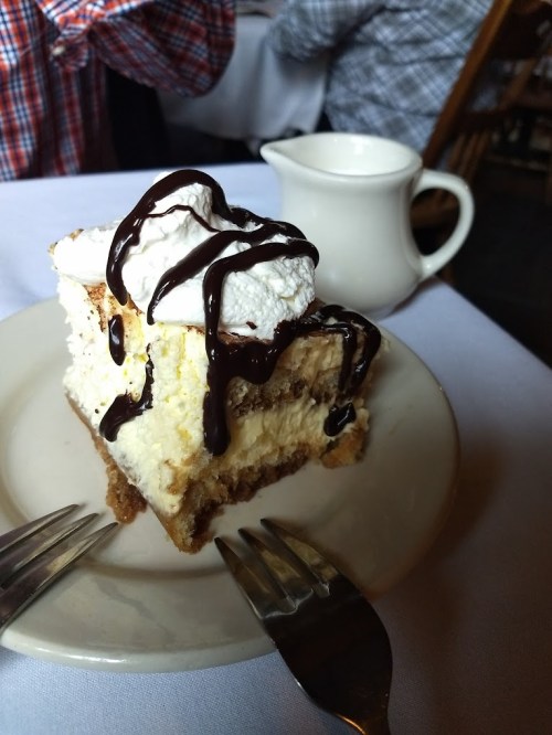 A slice of layered cheesecake topped with whipped cream and chocolate drizzle, served on a plate with two forks.