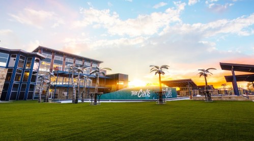 Modern building with palm trees and green lawn, set against a colorful sunset sky.