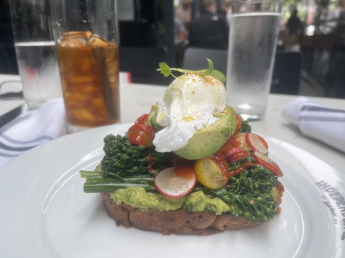 A plate with avocado toast topped with poached egg, cherry tomatoes, radishes, and greens, served in a restaurant setting.