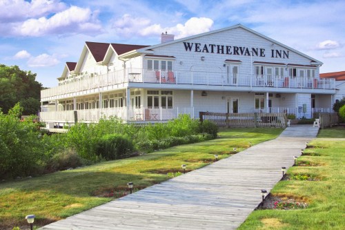 A white, two-story inn with a sign reading "Weathervane Inn," surrounded by green grass and a wooden pathway.