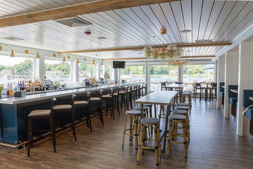 Bright, modern bar interior with wooden floors, high tables, and large windows overlooking a waterfront view.