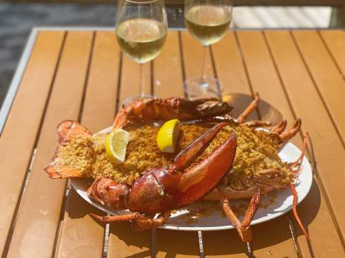 A plate of baked lobster topped with breadcrumbs, garnished with lemon, alongside two glasses of white wine.