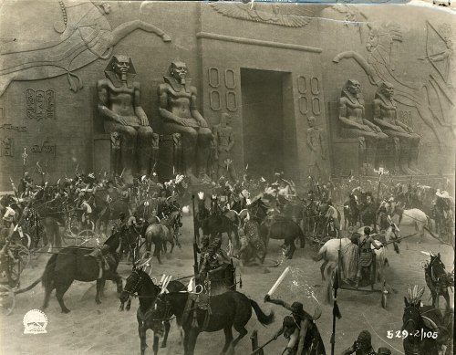 A historical scene depicting a large group of soldiers and horses near ancient statues and a grand entrance.