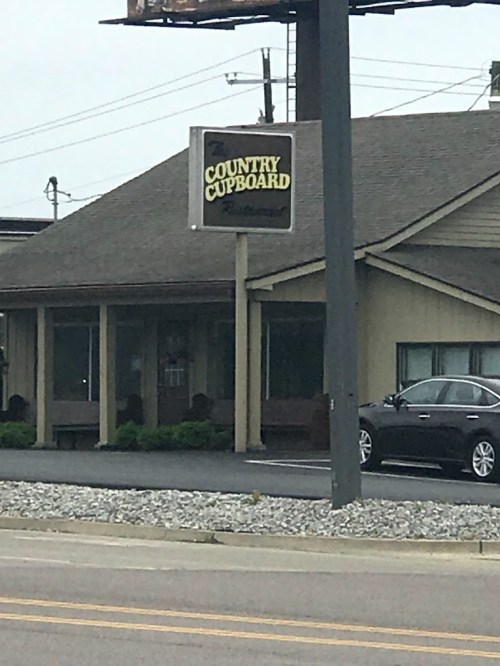 Sign for "Country Cupboard" restaurant with a building and parked car in the background, set along a roadside.