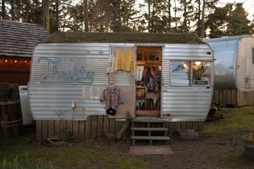 A vintage silver trailer with "Thrifty" sign, open door revealing clothes inside, surrounded by trees.