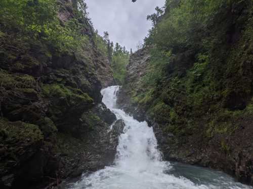 A cascading waterfall flows through rocky terrain, surrounded by lush green trees and a cloudy sky.