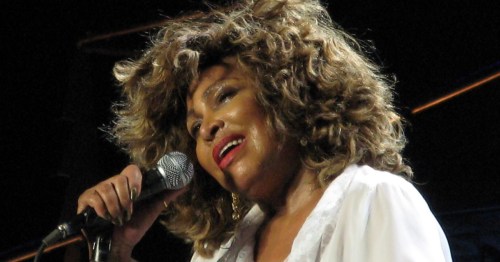 A singer with curly hair performs passionately on stage, holding a microphone and smiling.