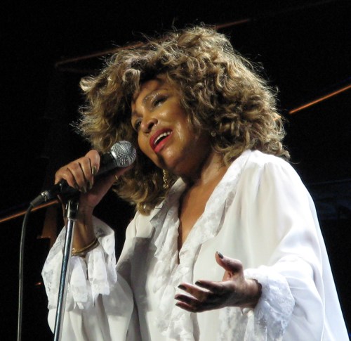 A singer with curly hair performs passionately on stage, holding a microphone and wearing a white outfit.