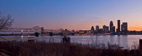City skyline at sunset with a bridge over a river, reflecting warm colors in the water and silhouetted buildings.