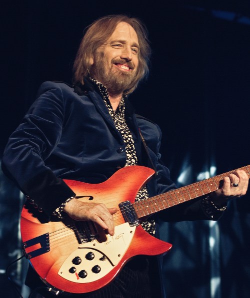 A musician smiles while playing an orange guitar on stage, wearing a blue velvet jacket and patterned shirt.