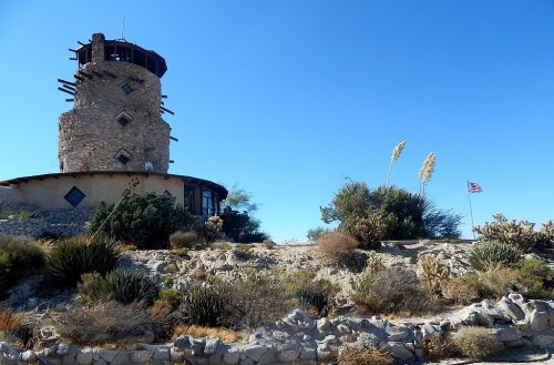 A stone tower with a wooden top stands on rocky terrain, surrounded by desert plants and an American flag in the background.