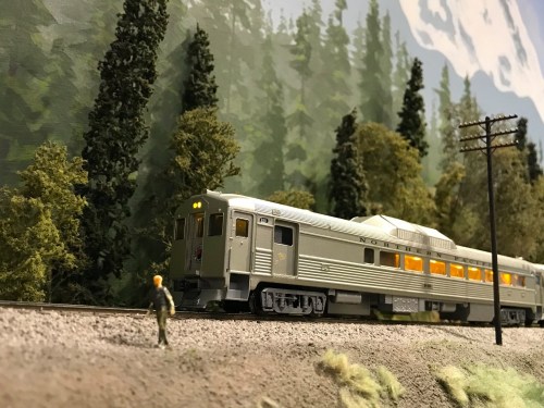 A model train passes through a scenic landscape with trees, while a miniature figure stands by the tracks.