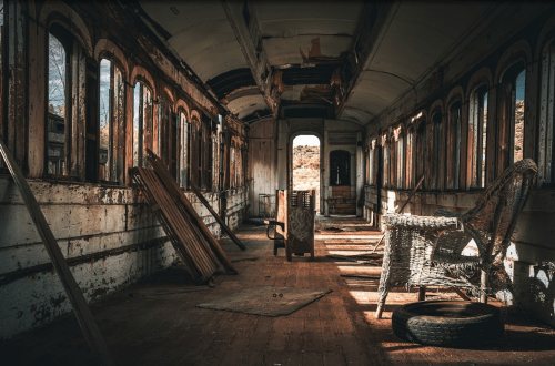 Abandoned train car interior with worn wooden benches, wicker chairs, and sunlight streaming through dusty windows.