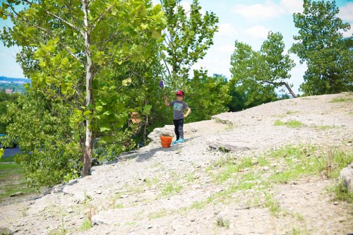 A child stands on a rocky slope, holding a purple object, with trees and a blue sky in the background.