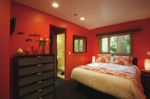 Cozy bedroom with red walls, floral bedding, and a bathroom door visible in the background. Natural light from a window.