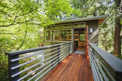 A modern cabin with large windows, surrounded by lush green trees, connected by a wooden walkway.