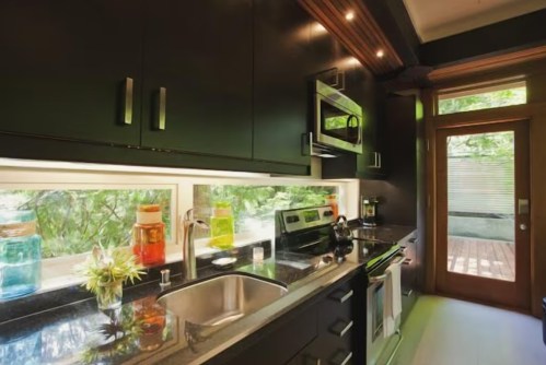 Modern kitchen with black cabinetry, stainless steel appliances, and colorful jars on the counter, featuring a view of greenery outside.