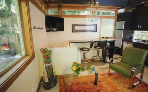A modern living space featuring a green chair, glass table, and bar stools, with large windows and colorful decor.