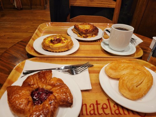 A wooden tray with pastries, a coffee cup, and forks on a table in a cozy café setting.