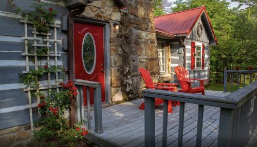 A cozy log cabin with a red door, stone accents, and two red chairs on a wooden porch surrounded by greenery.