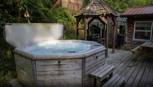 A wooden hot tub on a deck surrounded by greenery, with a gazebo and picnic table nearby.