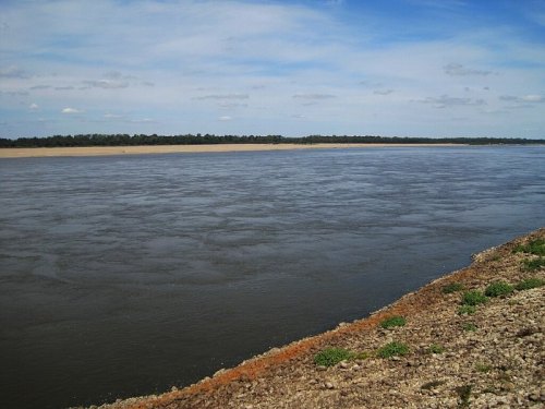 A calm river flows alongside a sandy bank under a blue sky with scattered clouds. Green vegetation lines the shore.