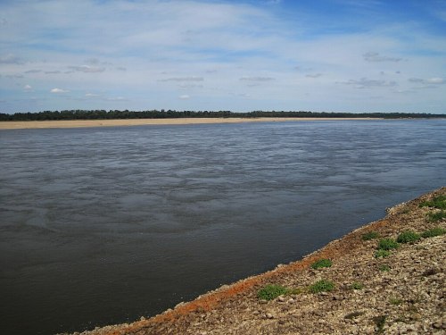 A calm river flows alongside a sandy bank under a blue sky with scattered clouds and greenery in the background.