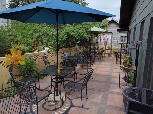 A patio with black metal tables and chairs, shaded by blue and green umbrellas, surrounded by greenery and flowers.