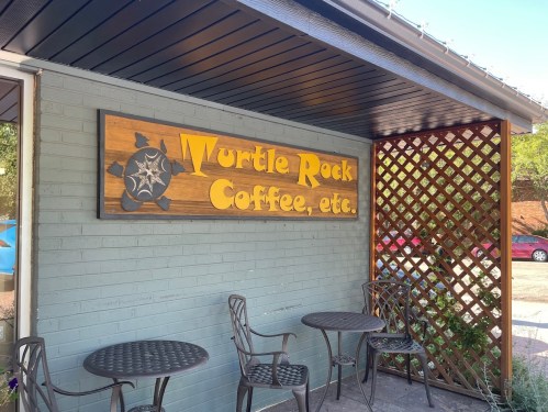 Sign for "Wurtle Rock Coffee, etc." on a blue wall, with two outdoor chairs and a lattice structure nearby.