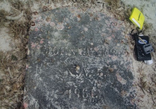 A weathered gravestone partially covered in sand, inscribed with "John Smith, N.Y., 1861," alongside a set of keys.