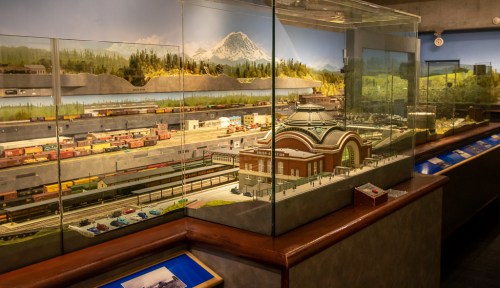 A detailed model train display featuring a scenic landscape, mountains, and a replica of a historic train station.