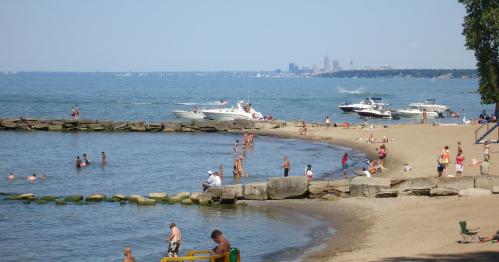 A sunny beach scene with people swimming and relaxing, boats on the water, and a city skyline in the background.