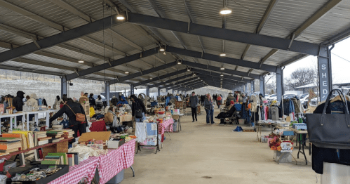 A bustling indoor market with vendors and shoppers browsing tables filled with various goods under a metal roof.