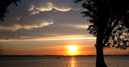 A serene sunset over a lake, with dramatic clouds and silhouettes of trees framing the scene.
